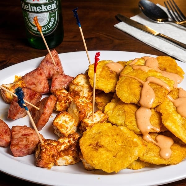 A plate of fried plantains with sauce, fried cheese, and sausage, with a Heineken bottle.